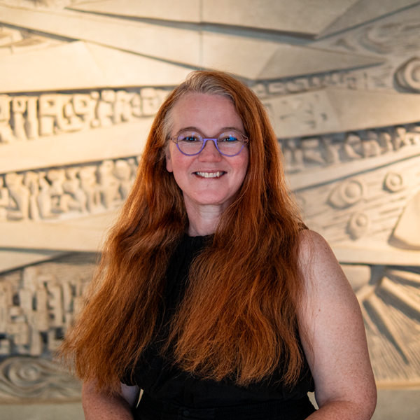 Head and shoulders portrait of Rebecca, wearing a sblack dress, standing in front of a textured light-coloured background.